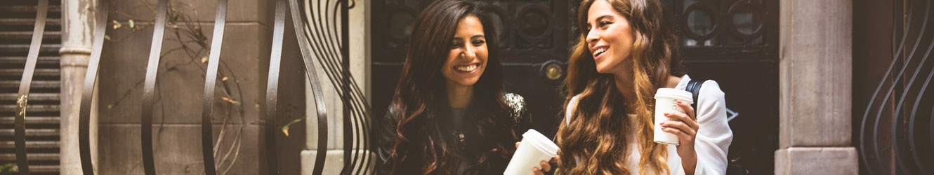 Two young women sitting together and laughing on a set of stairs. They are holding coffee.